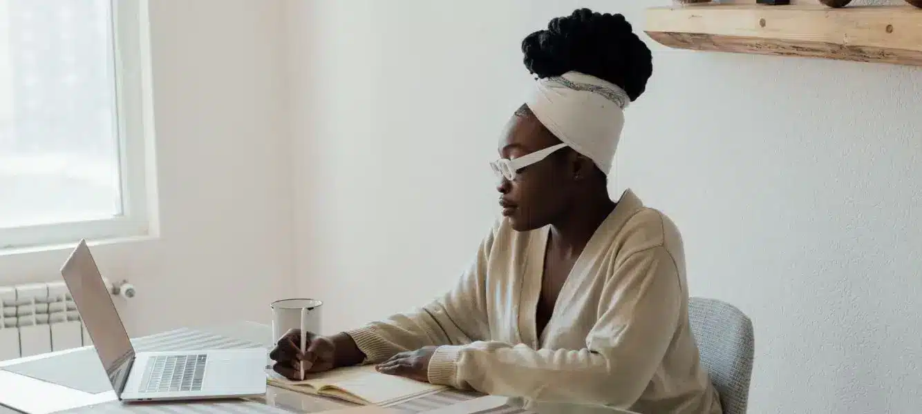 a woman is sat at a table, she is writing in a book and also looking at a laptop for the blog about flexible working